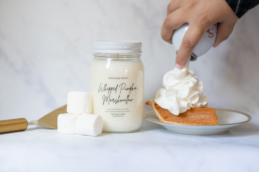 Jar of "whipped pumpkin marshmallow" scented candle with a hand dispensing whipped cream on top of a slice of pumpkin pie.