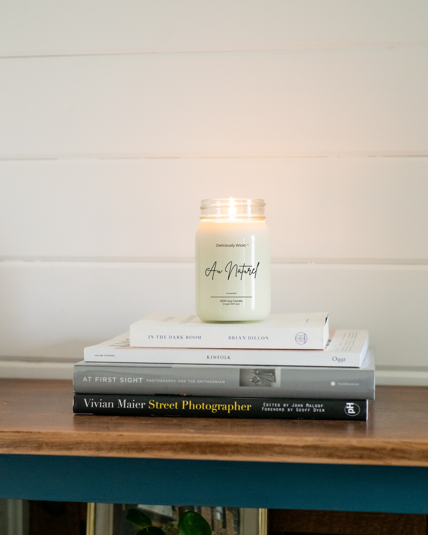 Candle labeled 'Au Naturel' on a stack of books with a white wall background