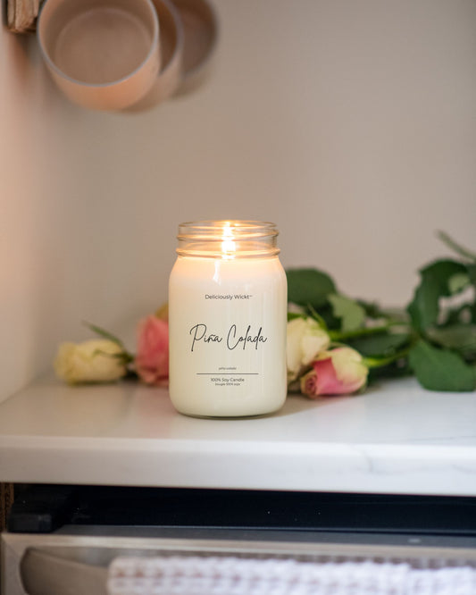 Deliciously Wickt Pina Colada 16 oz soy candle lit beside pink and white roses on a shelf.