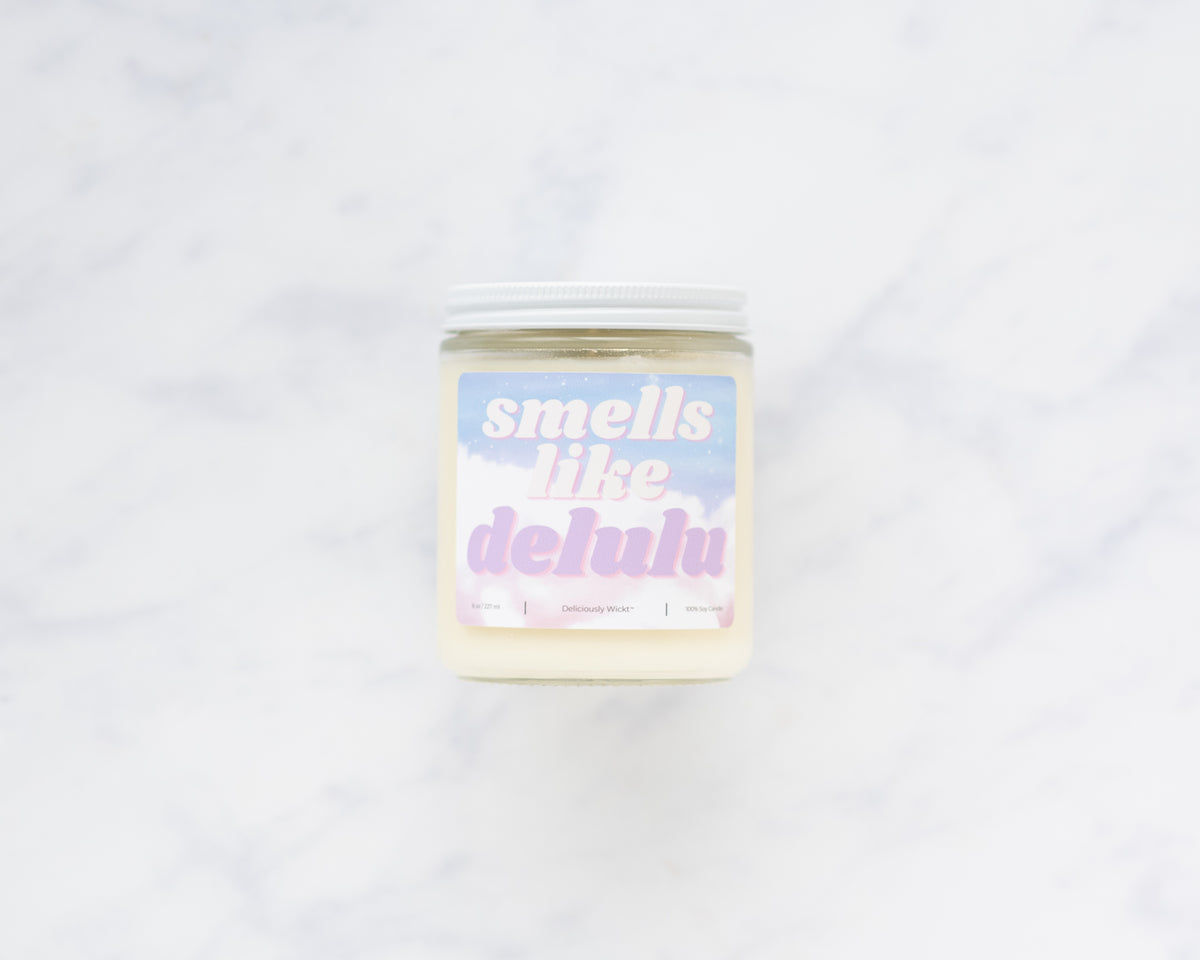 Deliciously Wickt Smells Like Delulu 8 oz soy candle with a white lid on a white marble surface.