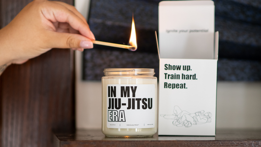 Deliciously Wickt In My Jiu-Jitsu Era 8 oz soy candle being lit with a match beside its "Show up. Train hard. Repeat." branded box.
