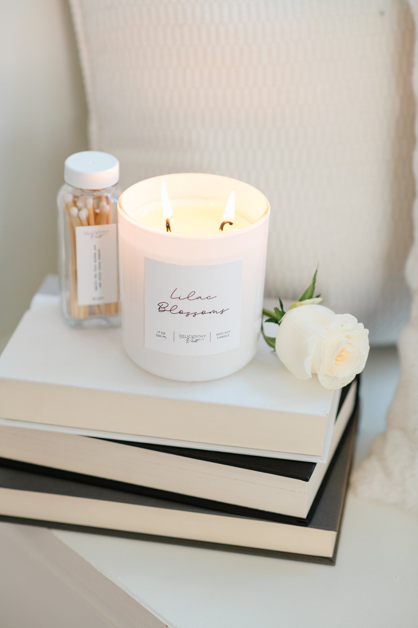 Deliciously Wickt glossy white 17 oz soy candle lit on stacked books beside a small bottle.