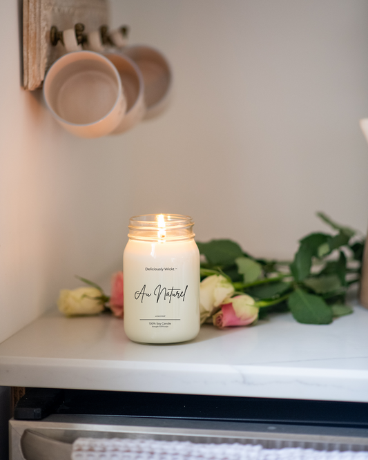 Candle labeled 'Au Naturel' on a shelf with flowers and mugs in the background