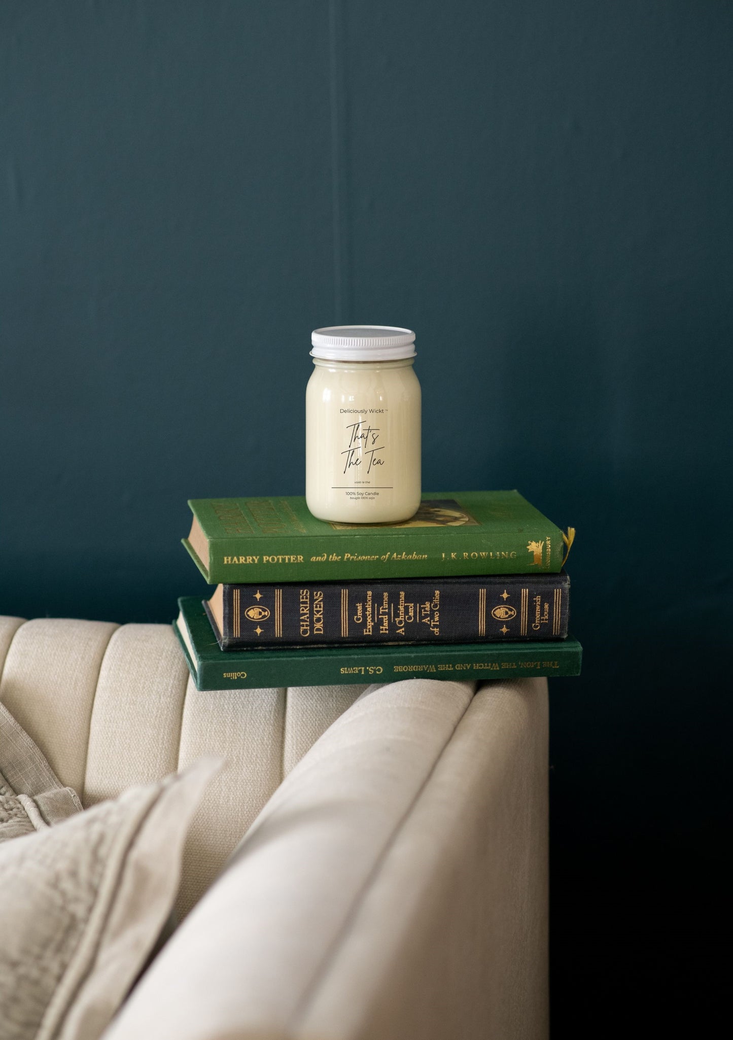 Mason jar candle scented "That's The tea" on top of books against a dark blue wall with a beige armchair
