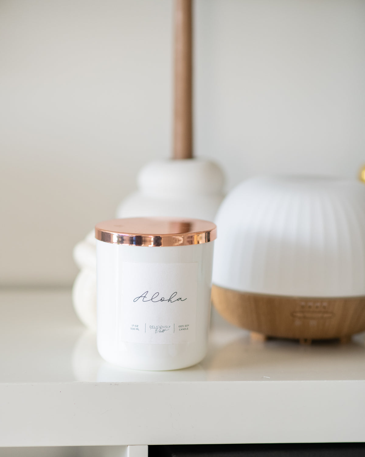 Deliciously Wickt Aloha 17 oz soy candle with rose gold lid displayed beside a white essential oil diffuser on a shelf.