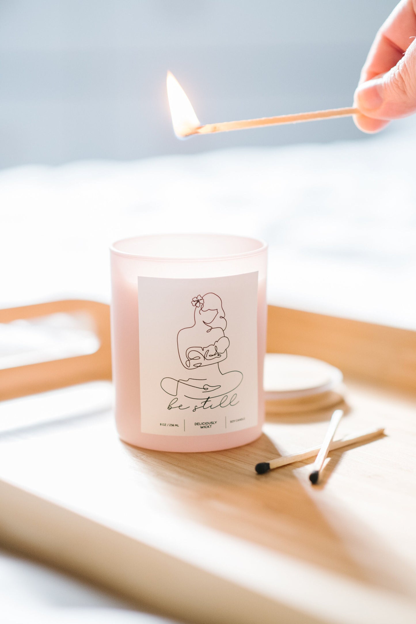 Deliciously Wickt Be Still dusty rose 8 oz soy candle being lit with a long stick match on a wooden tray.