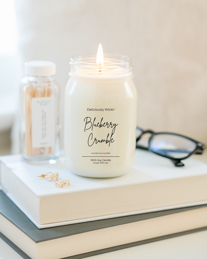 Lit Deliciously Wickt Blueberry Crumble 16 oz mason jar soy candle on stacked books with matches and black glasses.