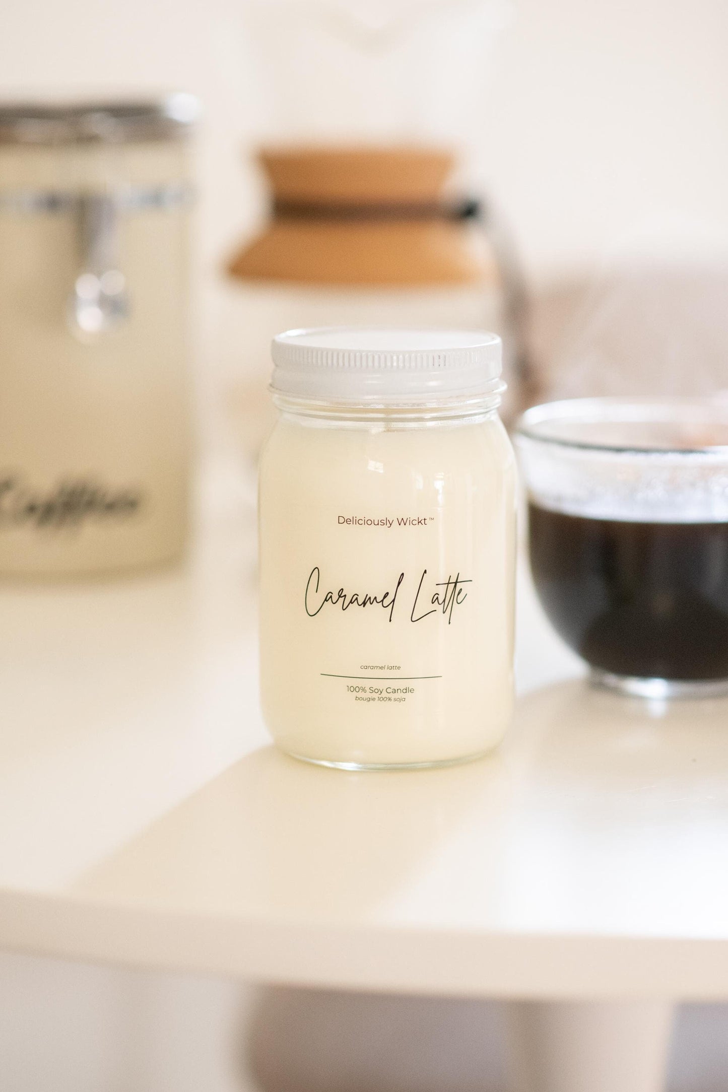Deliciously Wickt Caramel Latte 16 oz mason jar soy candle on a tabletop with a softly blurred neutral background.