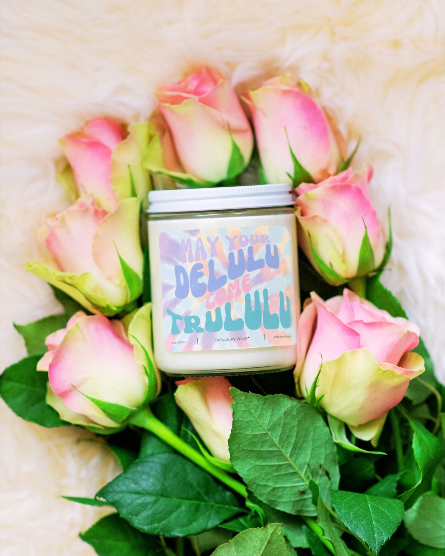 Deliciously Wickt Delulu Come Trululu 8 oz soy candle surrounded by pink roses on a soft white background.