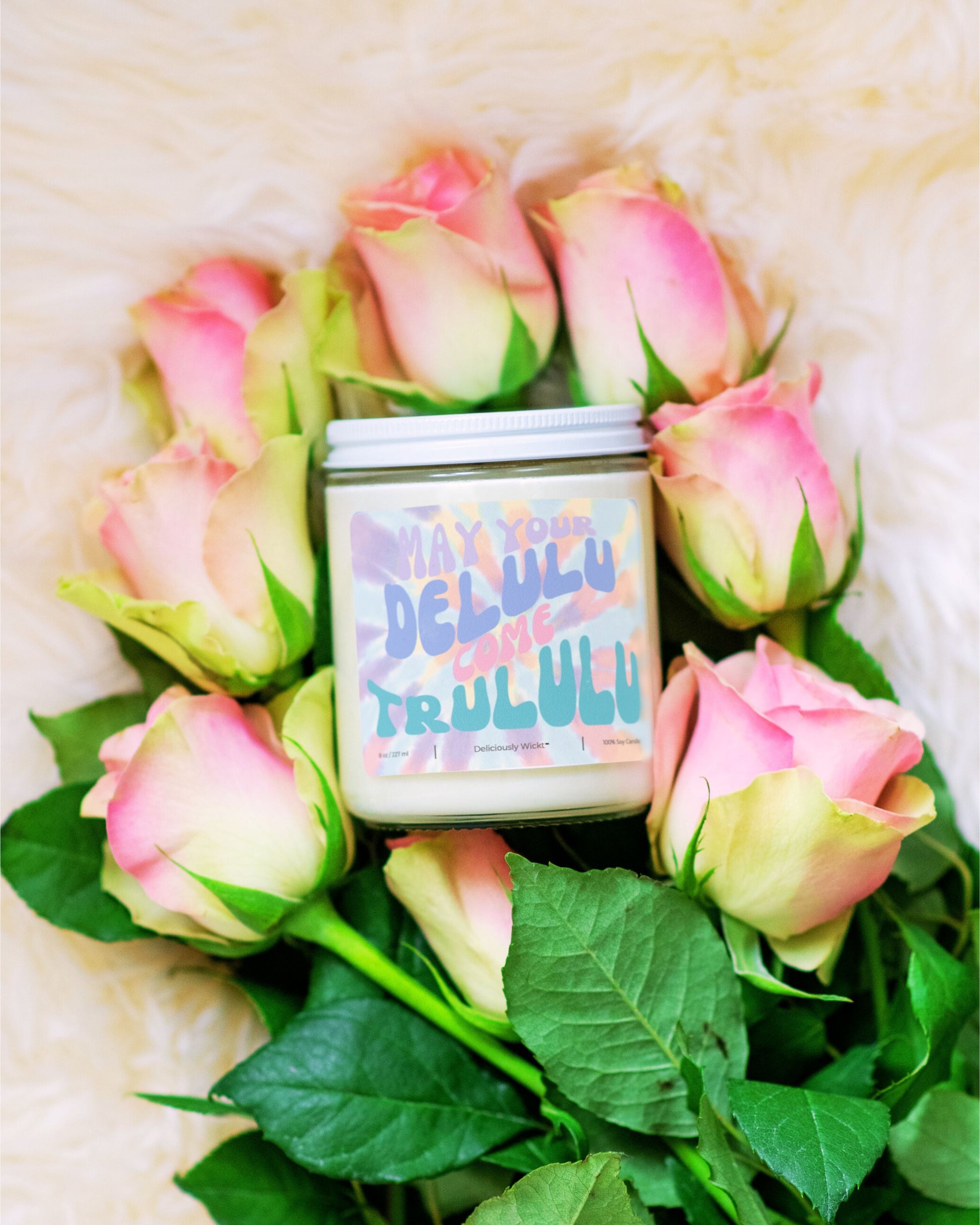 Deliciously Wickt Delulu Come Trululu 8 oz soy candle surrounded by pink roses on a soft white background.