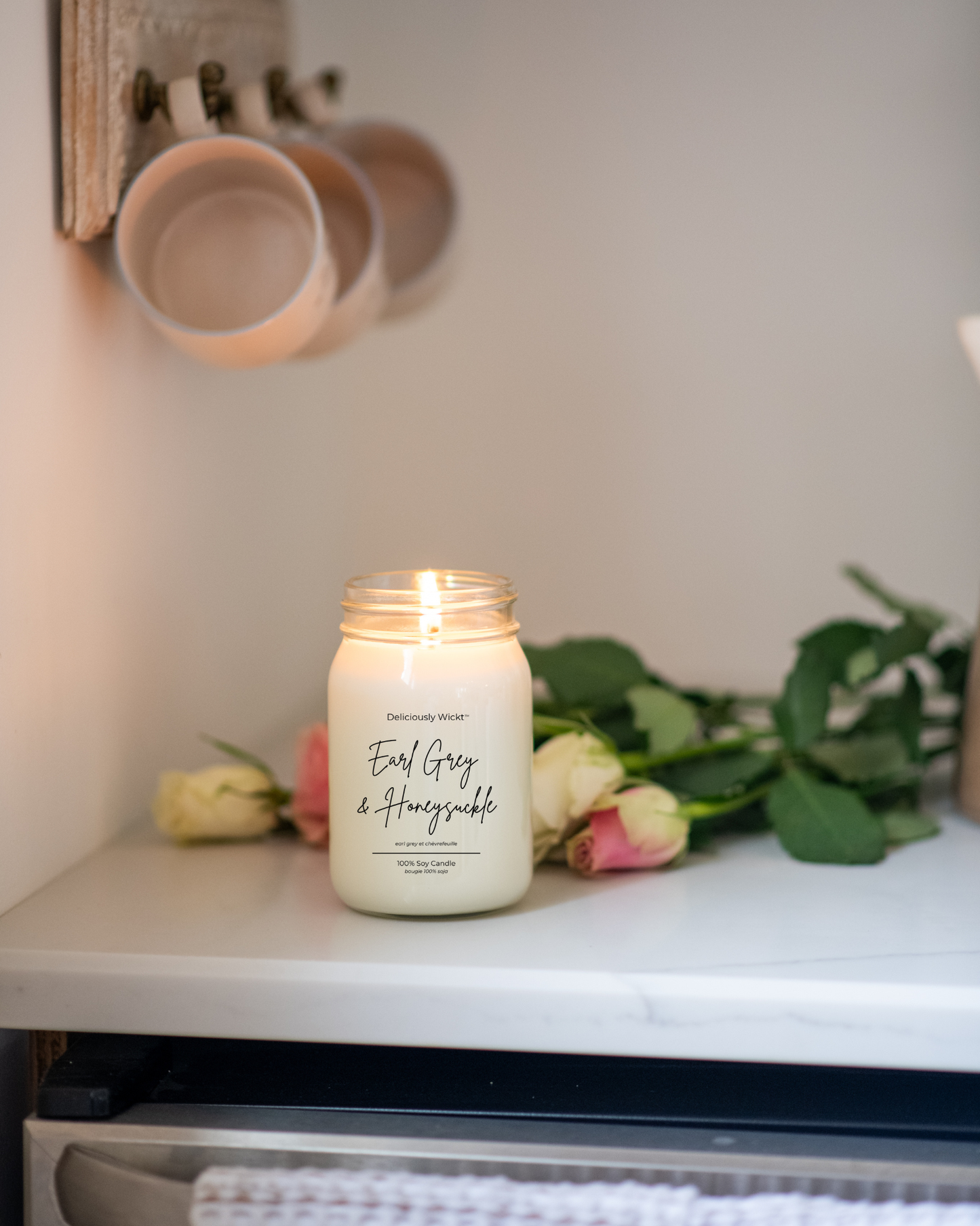 Lit Deliciously Wickt Earl Grey & Honeysuckle 16 oz soy candle on a white shelf with pink roses and hanging ceramic cups.
