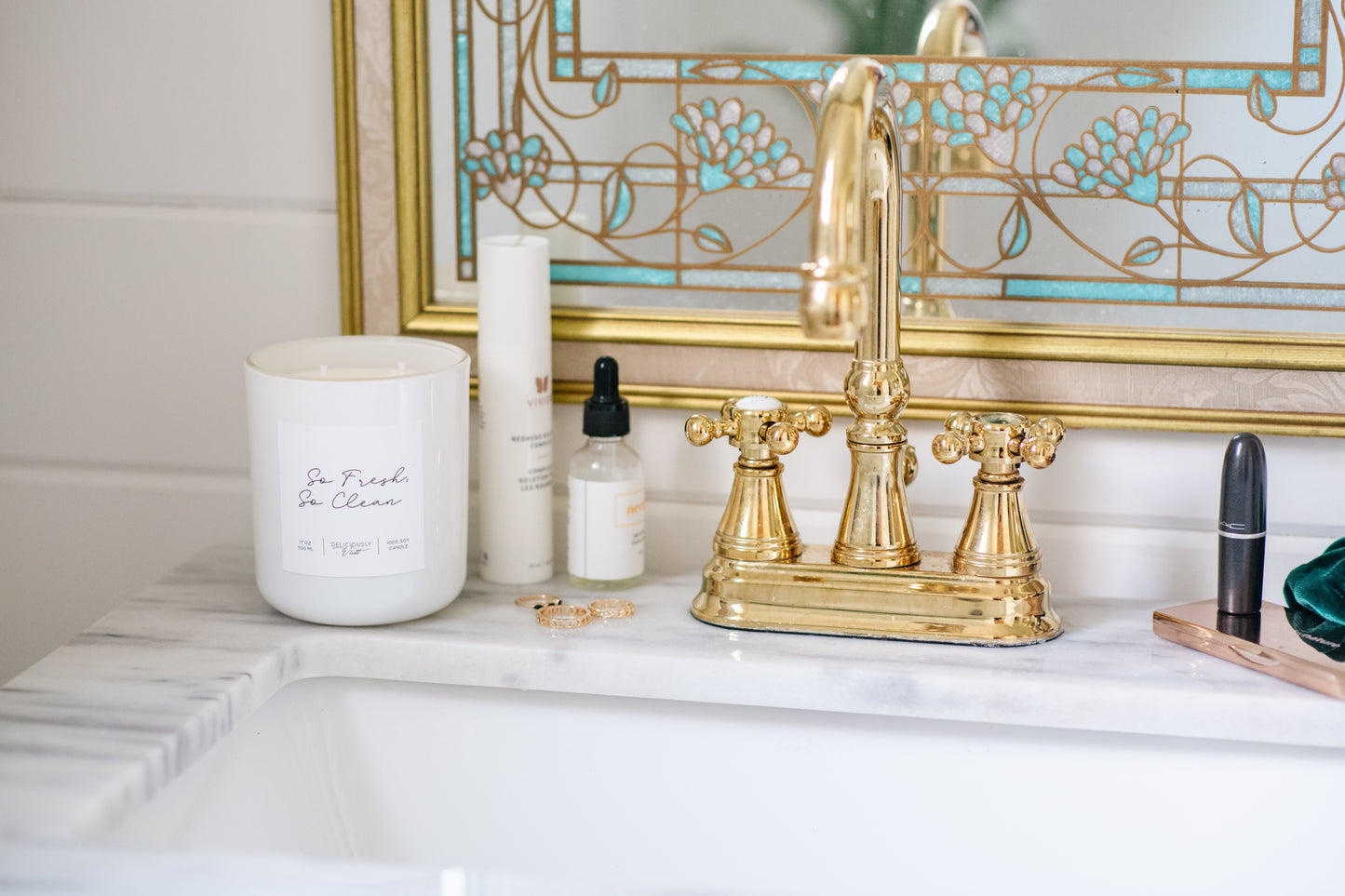 Deliciously Wickt glossy white 17 oz soy candle displayed beside gold fixtures on a bathroom counter.
