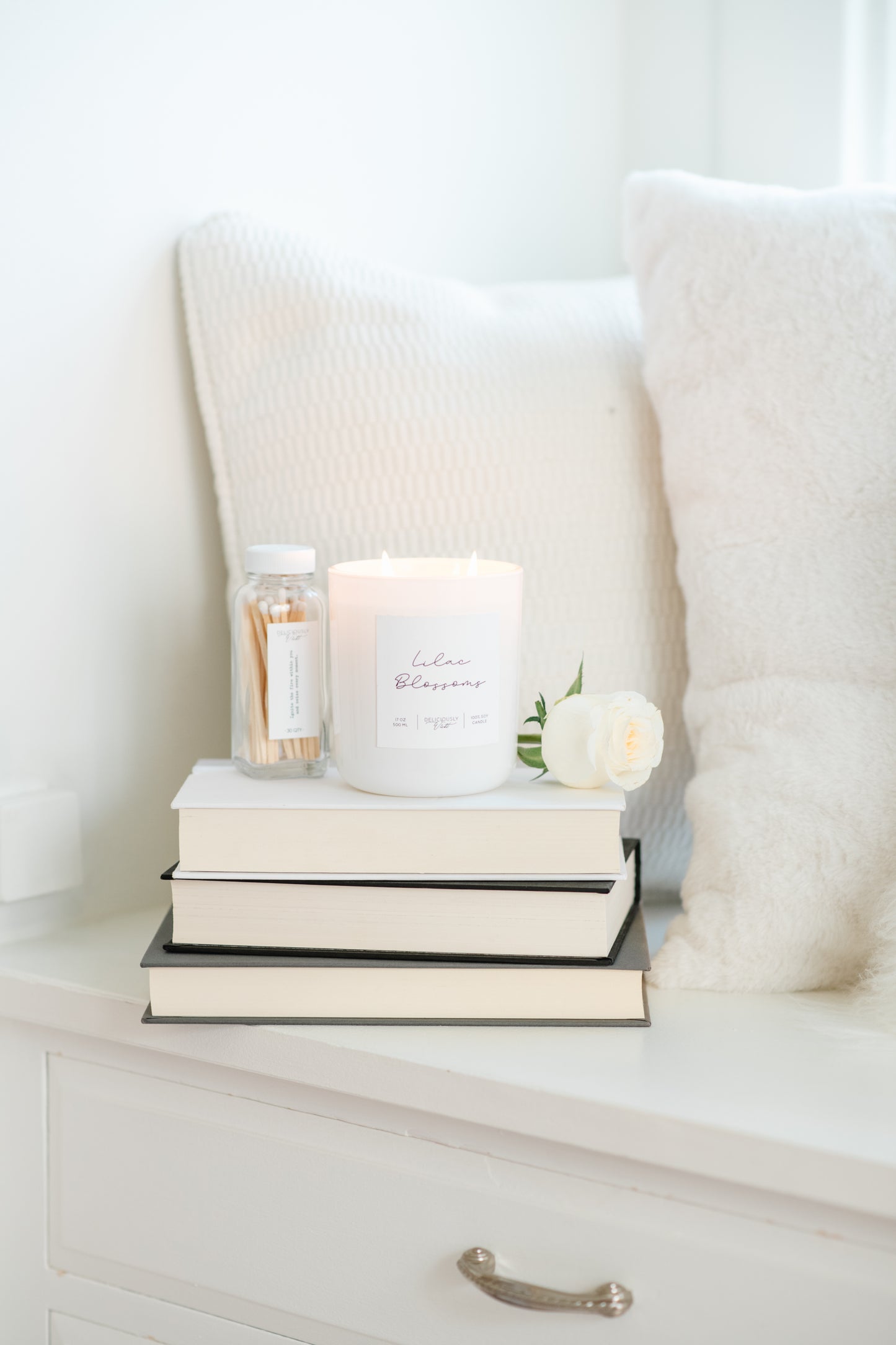 Deliciously Wickt glossy white 17 oz soy candle displayed on stacked books beside white bedding.