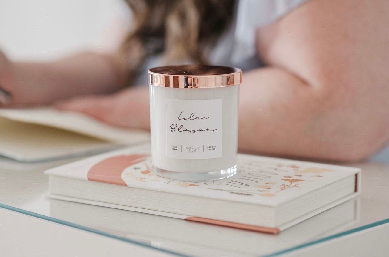 Deliciously Wickt glossy white 8 oz soy candle being lit while displayed on stacked books.
