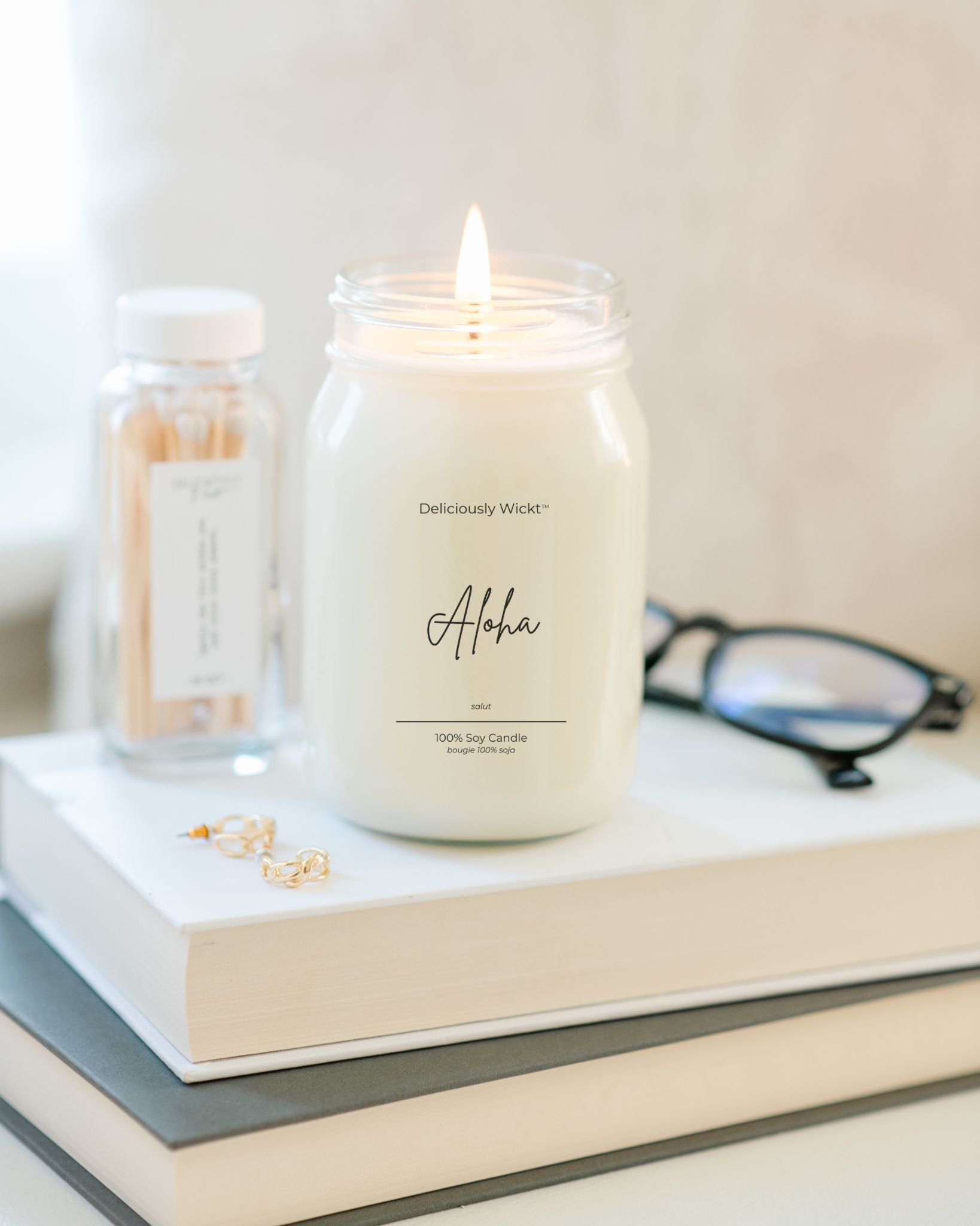 Deliciously Wickt Going, Going, Gone 16 oz mason jar soy candle lit on stacked books beside eyeglasses.