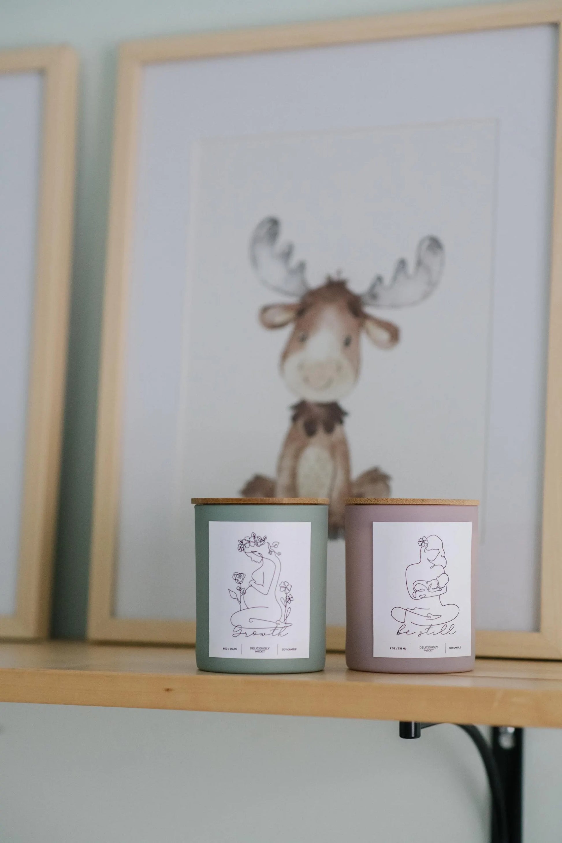 Deliciously Wickt Growth sage green and Be Still dusty rose 8 oz soy candles displayed together on a wooden nursery shelf beneath framed moose artwork.
