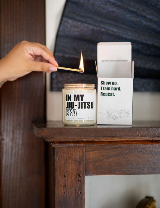 Deliciously Wickt In My Jiu-Jitsu Era 8 oz soy candle being lit on a wooden table beside its product box.