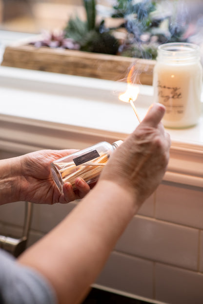 Lit Deliciously Wickt long stick safety match held beside a clear glass match bottle near a window.