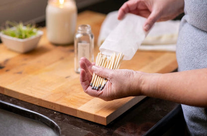 Deliciously Wickt long stick safety match refills being placed into a clear glass match bottle on a wooden counter.