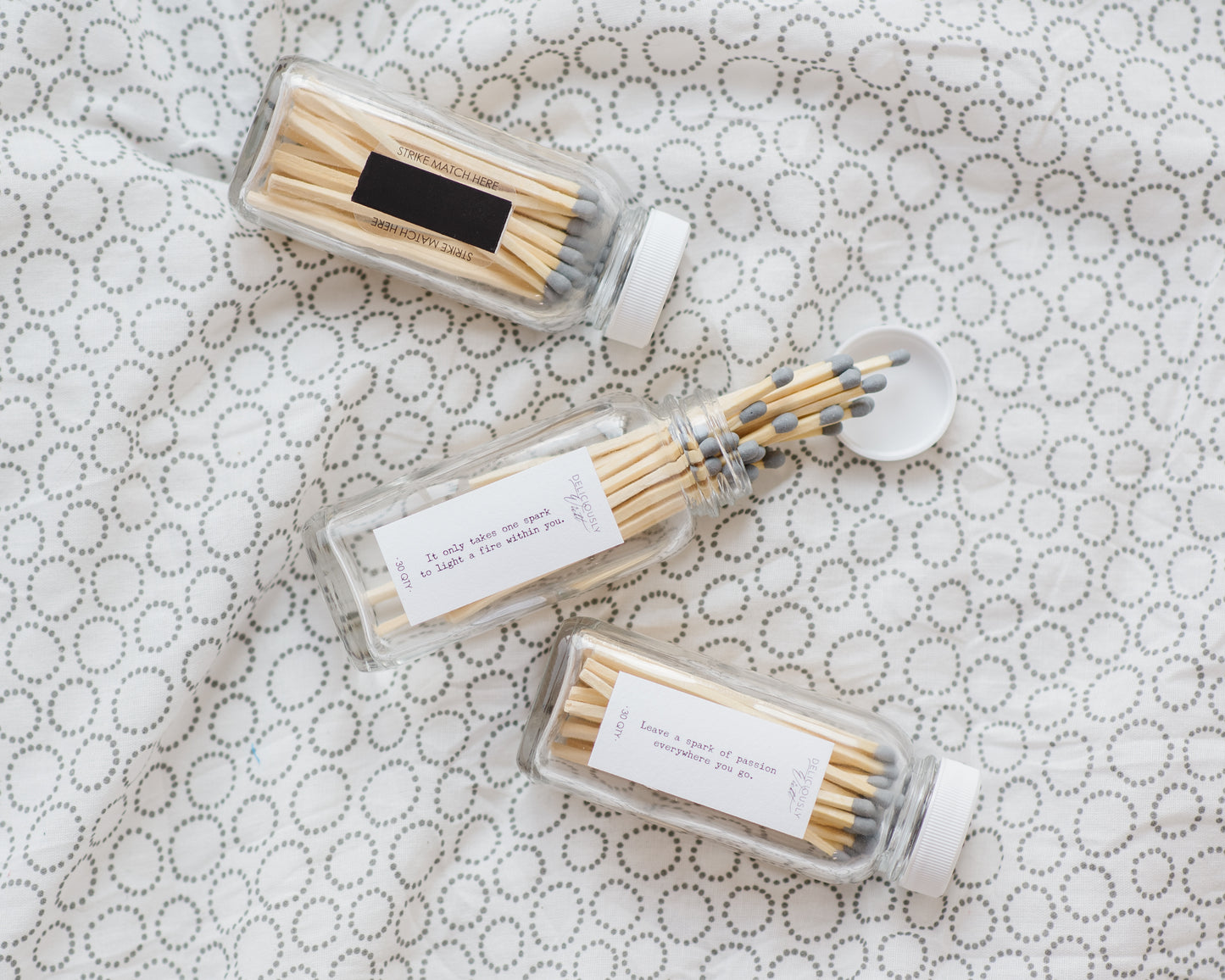 Three Deliciously Wickt glass match bottles with assorted long stick safety matches displayed on patterned fabric.