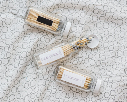 Three Deliciously Wickt glass match bottles with assorted long stick safety matches displayed on patterned fabric.