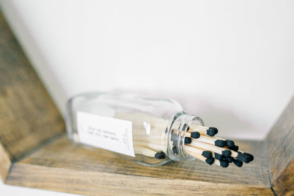 Deliciously Wickt long stick safety matches with black tips spilling from a clear glass match bottle on a wooden surface.