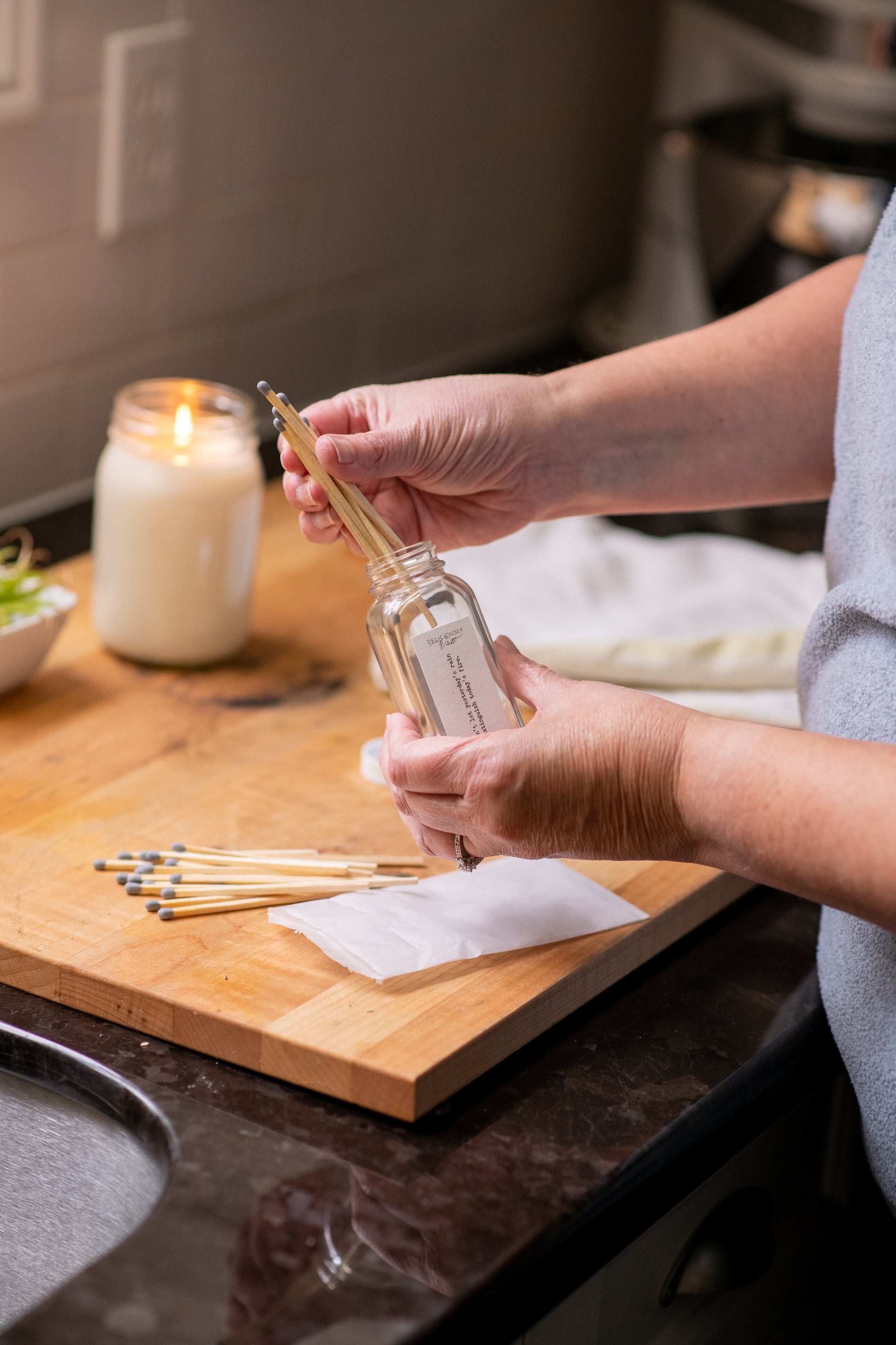 Deliciously Wickt long stick safety matches in a glass bottle being handled beside a lit soy candle on a wooden counter.