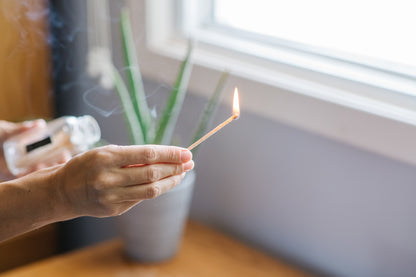 Deliciously Wickt long stick safety match burning beside a clear glass match bottle and houseplant.