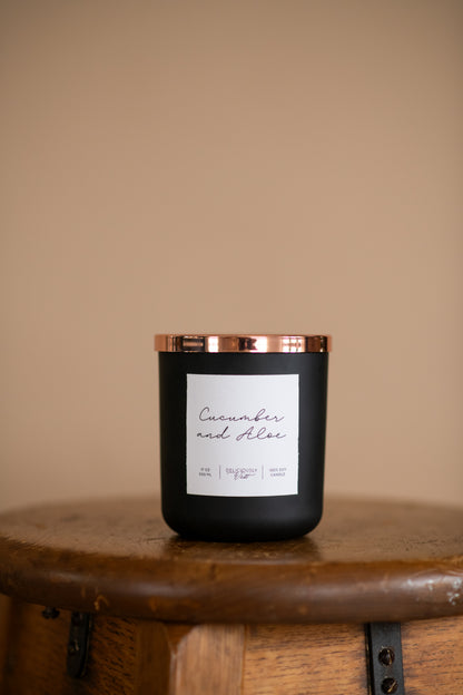 Deliciously Wickt matte black 17 oz soy candle displayed on a bathroom counter beside gold fixtures.
