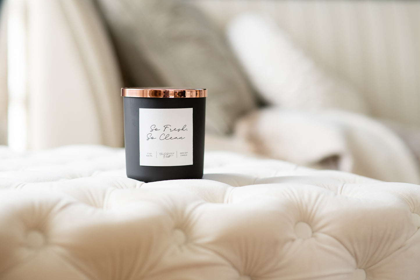 Deliciously Wickt matte black 17 oz soy candle displayed on soft white bedding.
