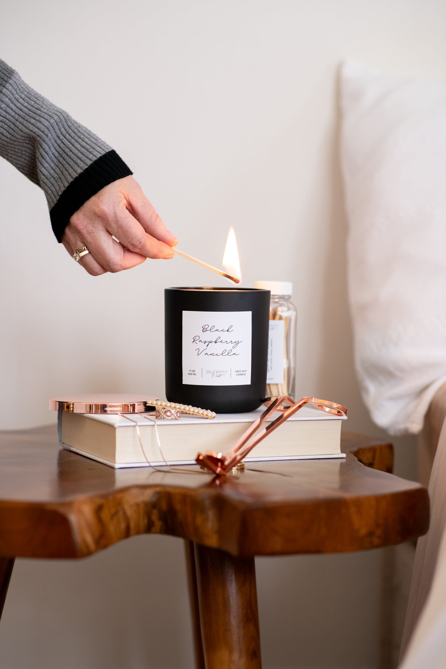 Deliciously Wickt matte black 17 oz soy candle being placed on a wooden bedside table beside white bedding.