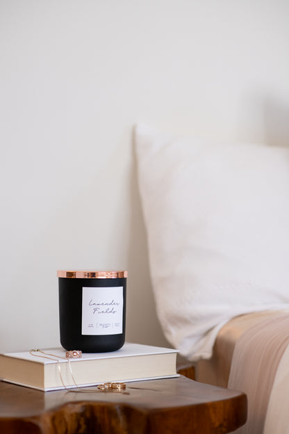 Deliciously Wickt matte black 17 oz soy candle with rose gold lid displayed on a wooden bedside table beside white bedding.