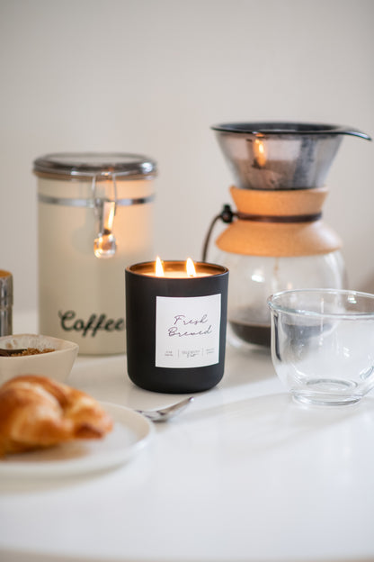 Deliciously Wickt matte black 17 oz soy candle styled on a table beside coffee and pastries.