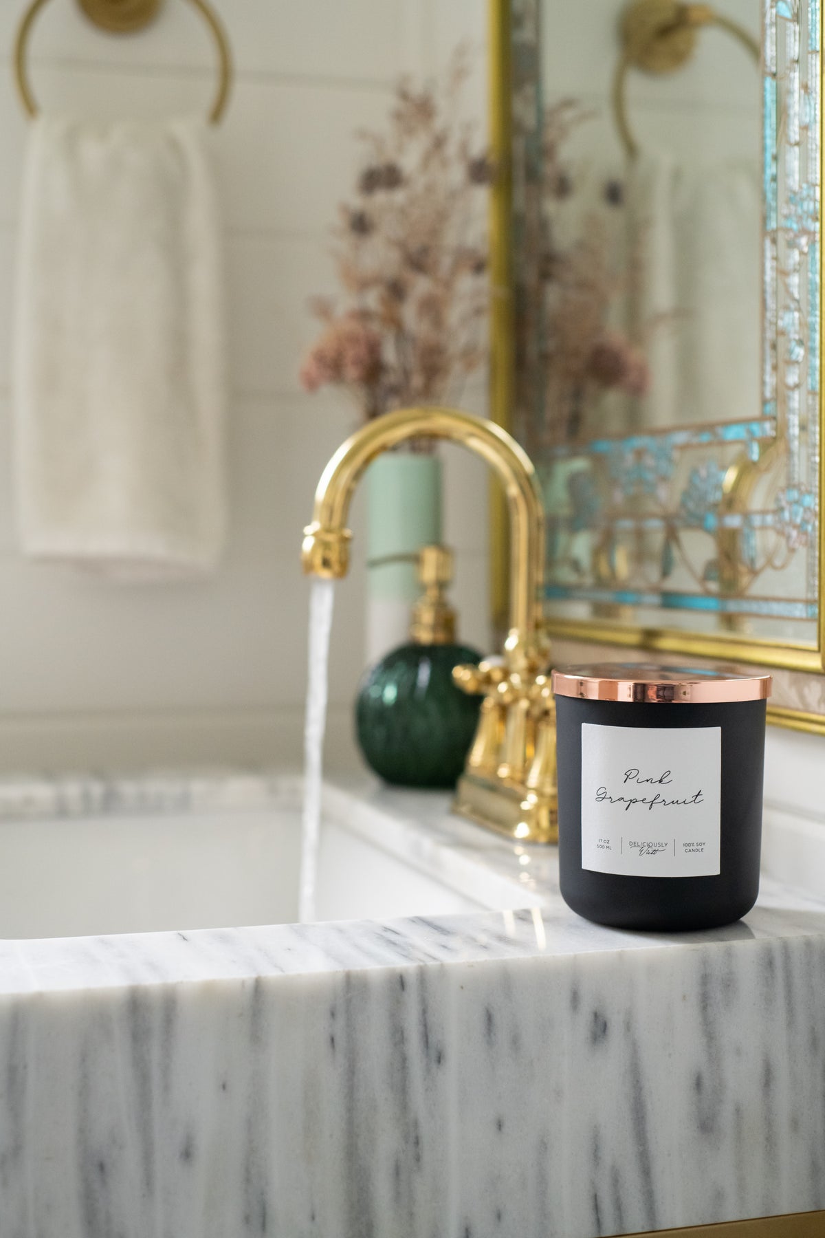 Deliciously Wickt matte black 17 oz soy candle with rose gold lid on a marble bathroom sink beside a gold faucet.