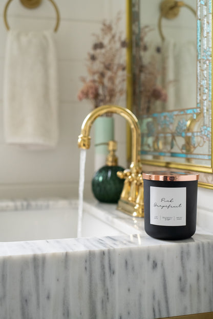 Deliciously Wickt matte black 17 oz soy candle with rose gold lid on a marble bathroom sink beside a gold faucet.