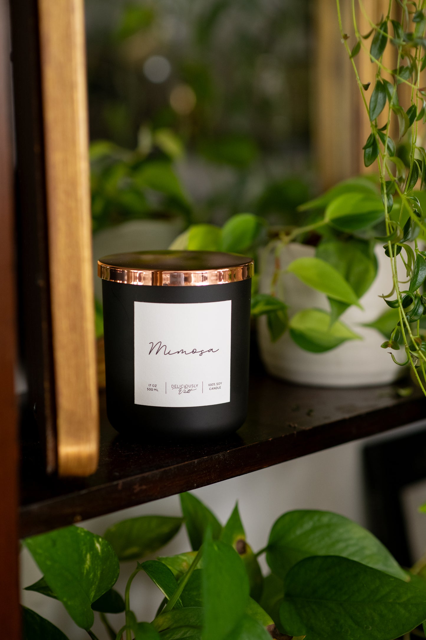 Deliciously Wickt matte black 17 oz soy candle displayed among green houseplants.