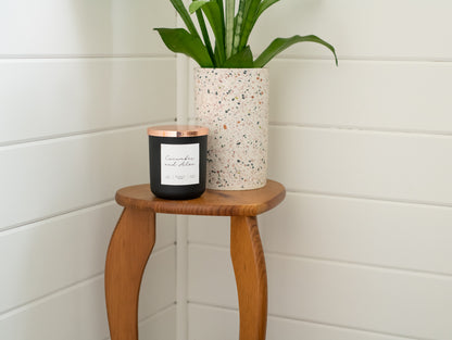 Deliciously Wickt matte black 17 oz soy candle displayed on a small wooden stool beside a green plant.