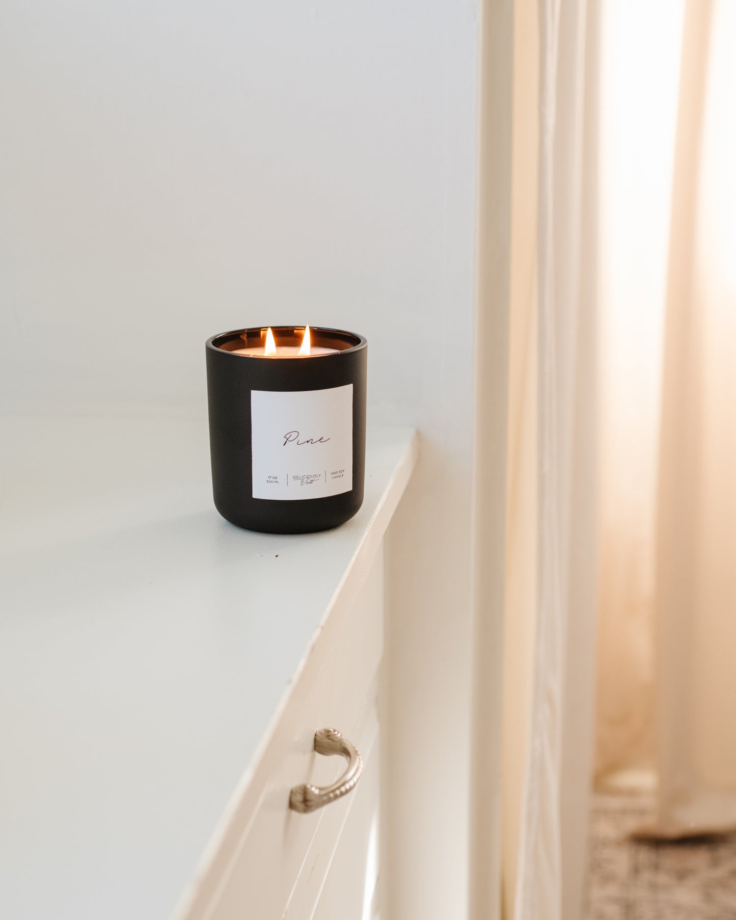 Deliciously Wickt matte black 17 oz soy candle displayed on a white ledge beside a sheer curtain