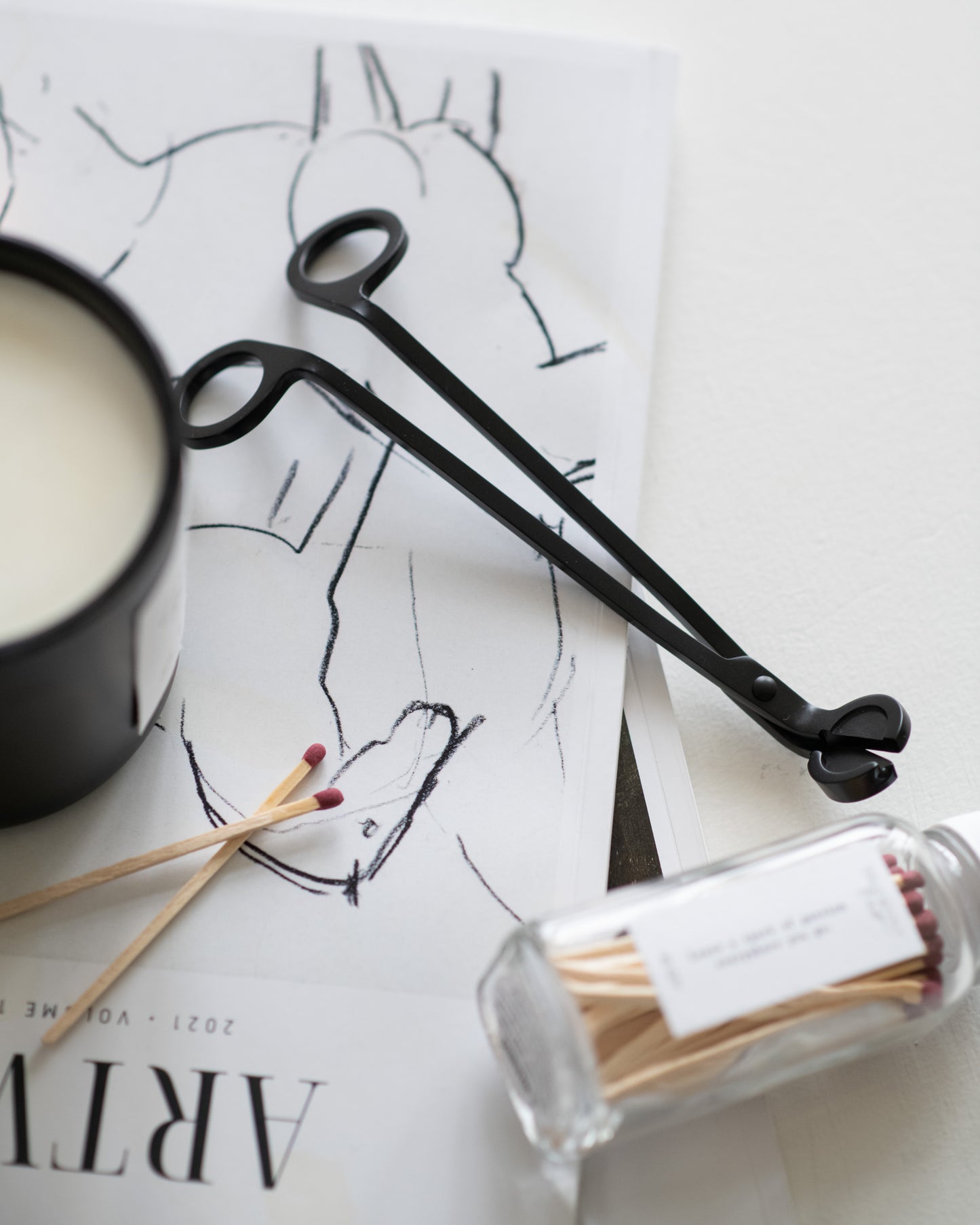 Deliciously Wickt black metal candle wick trimmer displayed beside a soy candle and glass bottle of long stick matches.