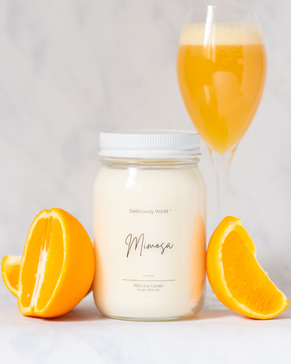 Deliciously Wickt Mimosa 16 oz mason jar soy candle displayed with fresh orange halves and a glass of sparkling orange mimosa.