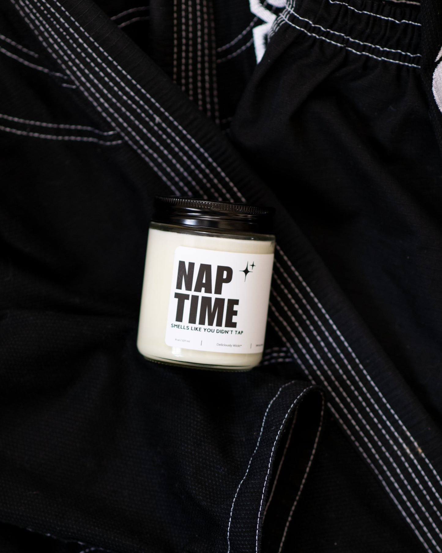Nap Time Jiu-Jitsu soy candle with “Smells like you didn’t tap” label displayed on a black gi