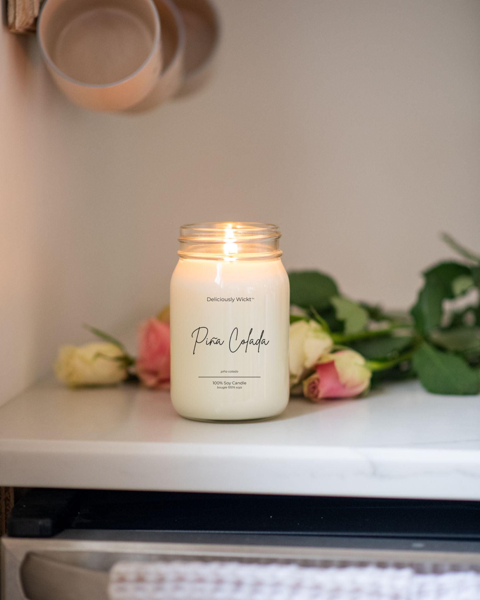 Deliciously Wickt Pina Colada 16 oz soy candle lit beside pink and white roses on a shelf.