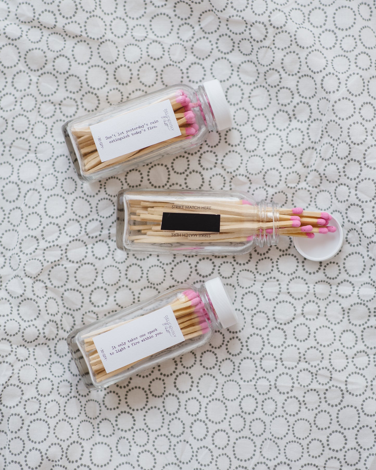 Three Deliciously Wickt glass bottles filled with pink-tipped long stick safety matches on patterned fabric.