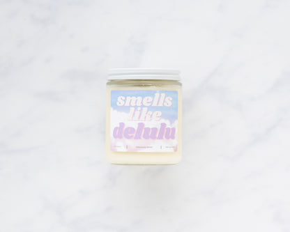 Deliciously Wickt Smells Like Delulu 8 oz soy candle with a white lid on a white marble surface.