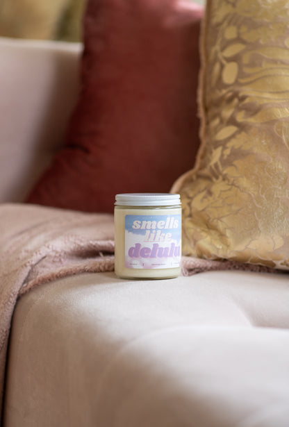 Deliciously Wickt Smells Like Delulu 8 oz soy candle on a pink sofa beside a rose and gold cushion.