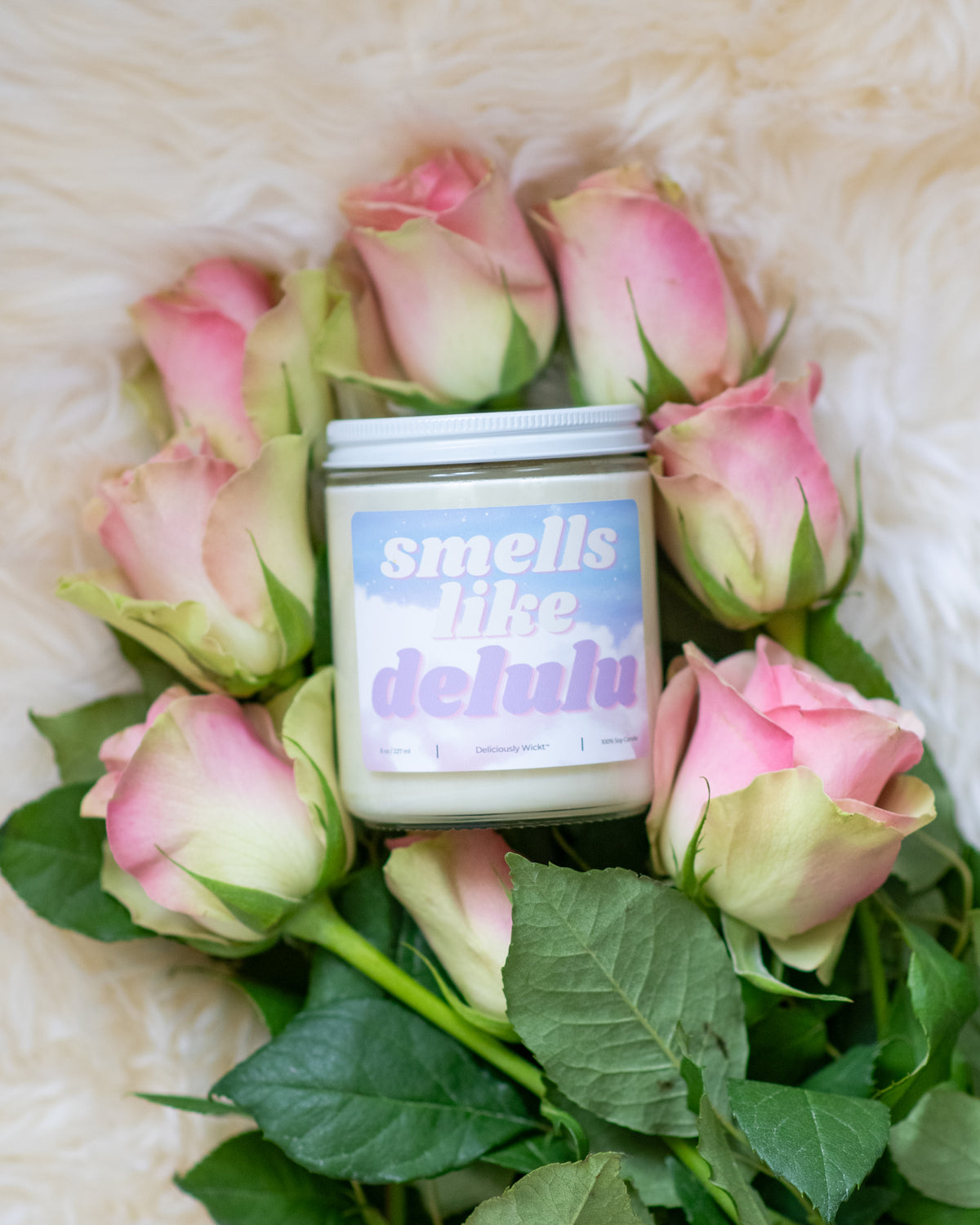 Deliciously Wickt Smells Like Delulu 8 oz soy candle surrounded by pink roses on a soft white rug.
