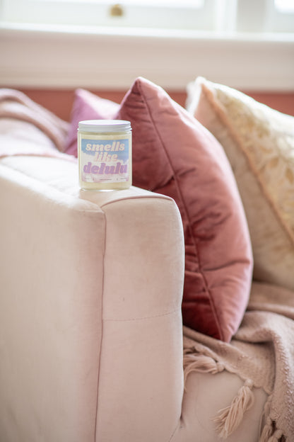 Deliciously Wickt Smells Like Delulu 8 oz soy candle on the arm of a pink sofa beside a rose velvet cushion.