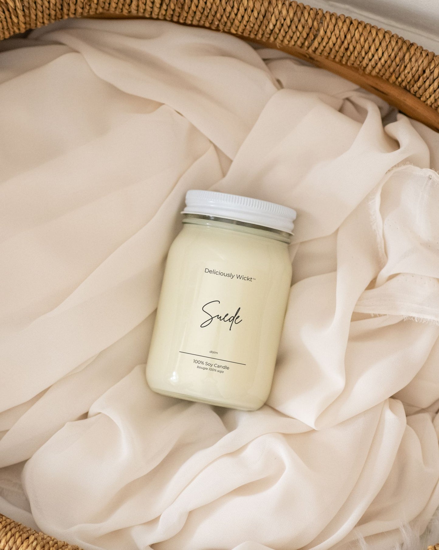 Deliciously Wickt Suede 16 oz soy candle resting on soft cream fabric in a woven basket.