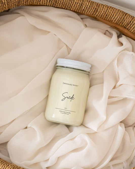 Deliciously Wickt Suede 16 oz soy candle resting on soft cream fabric in a woven basket.
