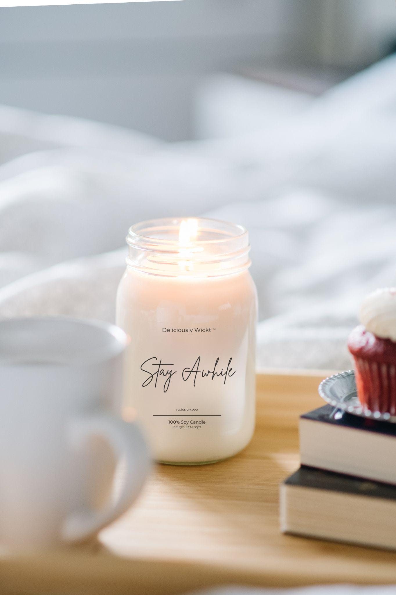 Candle labeled 'Stay Awhile' on a table with a cup, books, and a cupcake.